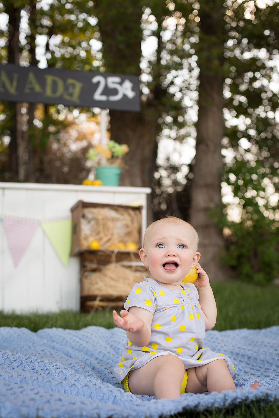 Little girl drops a lemon behind her back while sitting on a blanket in front of lemonade stand for her first birthday photos.