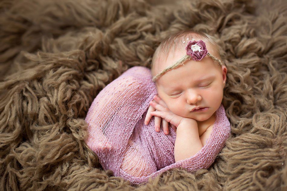 Baby girl wearing pink sleeps curled up on brown fur while being photographed by Pueblo Newborn photographer.