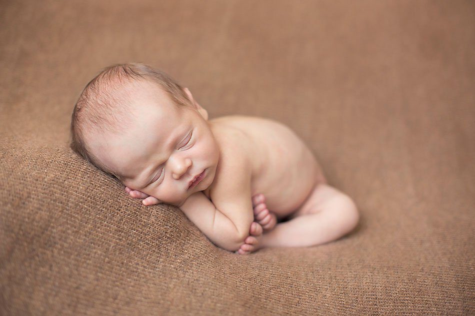 Baby boy sleeps curled up on a brown blanket during photo shoot with Pueblo newborn photographer.
