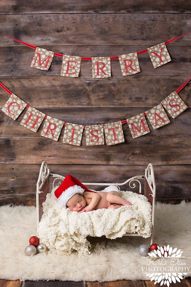 Baby boy wearing a Santa hat and sleeping on a tiny bed by Pueblo Newborn Photographer K.D. Elise Photography.