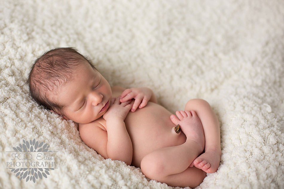 Newborn boy sleeps curled up with his arms on his chest on a cream blanket.  Newborn Photography by Pueblo CO photographer K.D. Elise Photography.