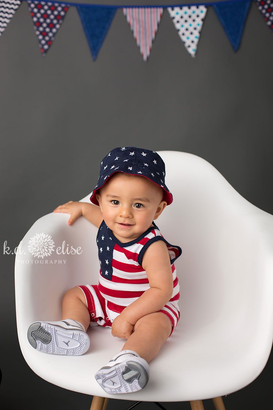 Baby boy in red white and blue outfit sitting in a white pyramid chair. Baby photography by Pueblo children's photographer K.D. Elise Photography.