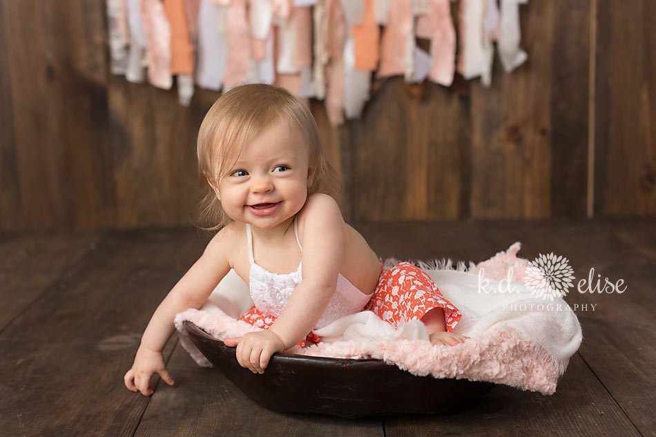 Smiling baby girl in orange outfit, smiling at the camera during her 9 month photoshoot with Pueblo photographer K.D. Elise Photography.