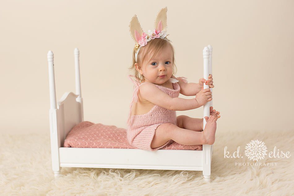 Little girl in a bunny outfit sitting on a white wooden baby bed during her nine month photoshoot with K.D. Elise Photography.