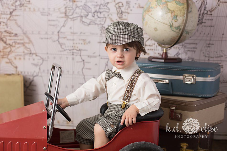 Little boy in a vintage peddle-car poses for portraits in front of a world map backdrop. Themed children's photoshoot by K.D. Elise Photography.