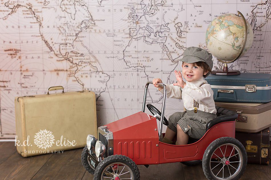 Little boy in vintage red peddle-car, waving at camera. Themed children's photoshoot by K.D. Elise Photography.