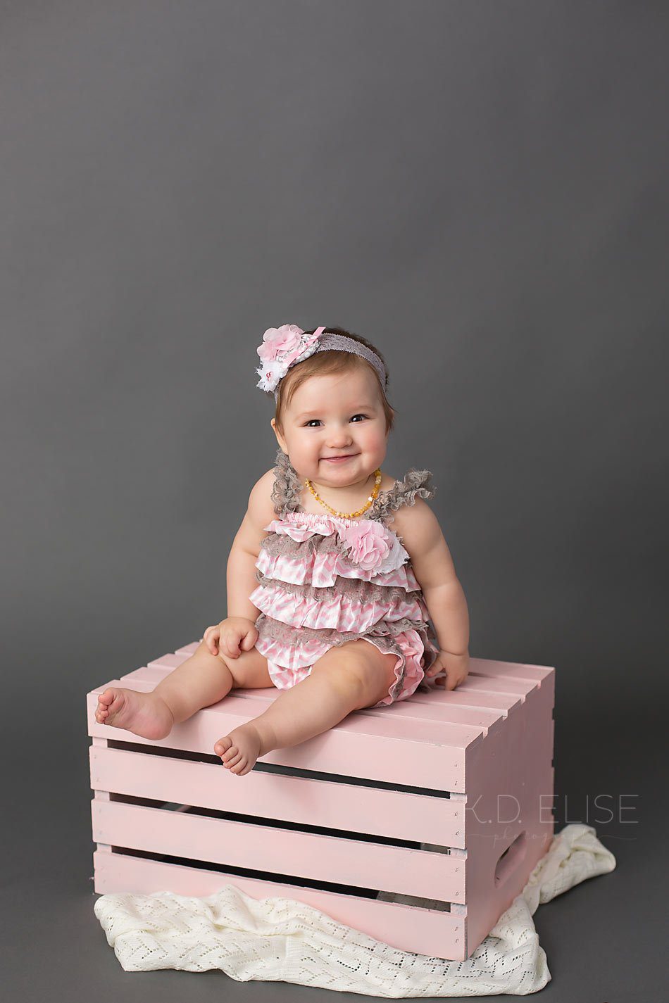 Smiling baby girl sitting on a pink crate in front of a grey backdrop. Colorado Springs baby photography by K.D. Elise Photography.