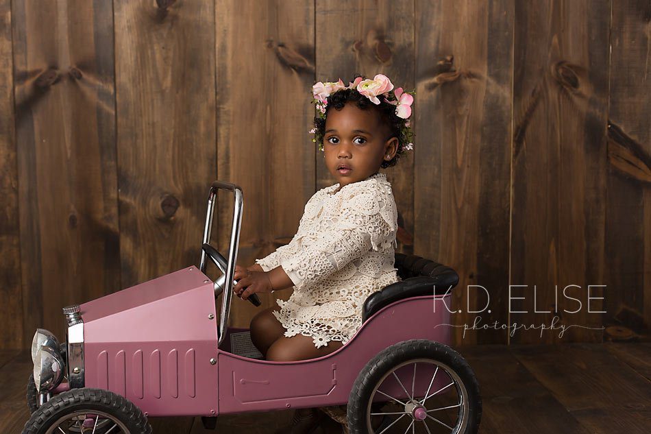 Little girl in cream dress and pink flower headband sitting in a pink peddle car. Photo by Pueblo children's photographer K.D. Elise Photography.