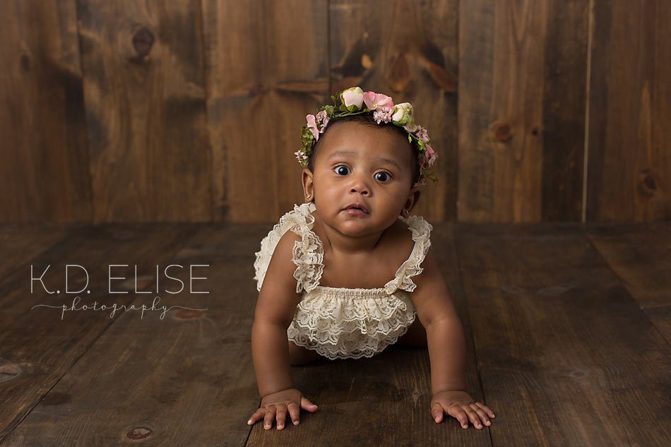 Baby girl in cream romper and floral headband crawling on a dark wood backdrop, by Pueblo children's photographer K.D. Elise Photography