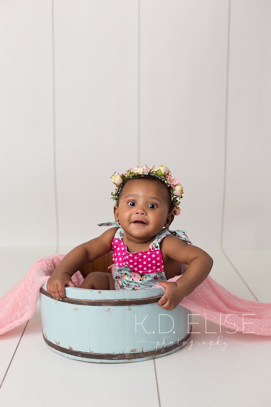 Baby girl in blue romper and floral headband sitting in a blue antique style bucket, by Pueblo children's photographer K.D. Elise Photography.