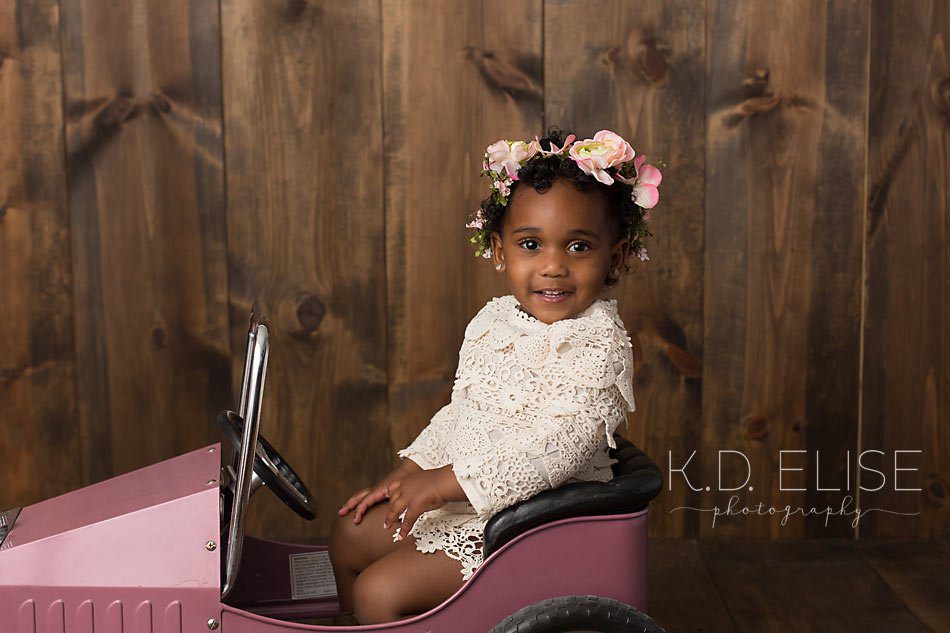 Little girl in cream dress and floral headband sits in a pink peddle car and smiles at the camera during photos with Pueblo children's photographer K.D. Elise Photography.