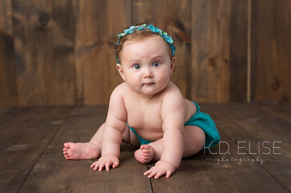 Baby girl in teal outfit sitting on a wooden backdrop. Baby portraits by Colorado Springs photographer K.D. Elise Photography.