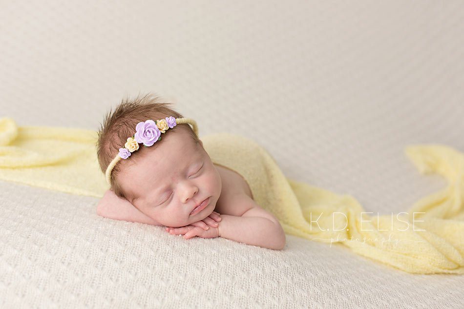 Newborn girl on cream blanket, facing the camera while wearing yellow wrap and purple and yellow headband. Newborn photography by Pueblo photographer K.D. Elise Photography.