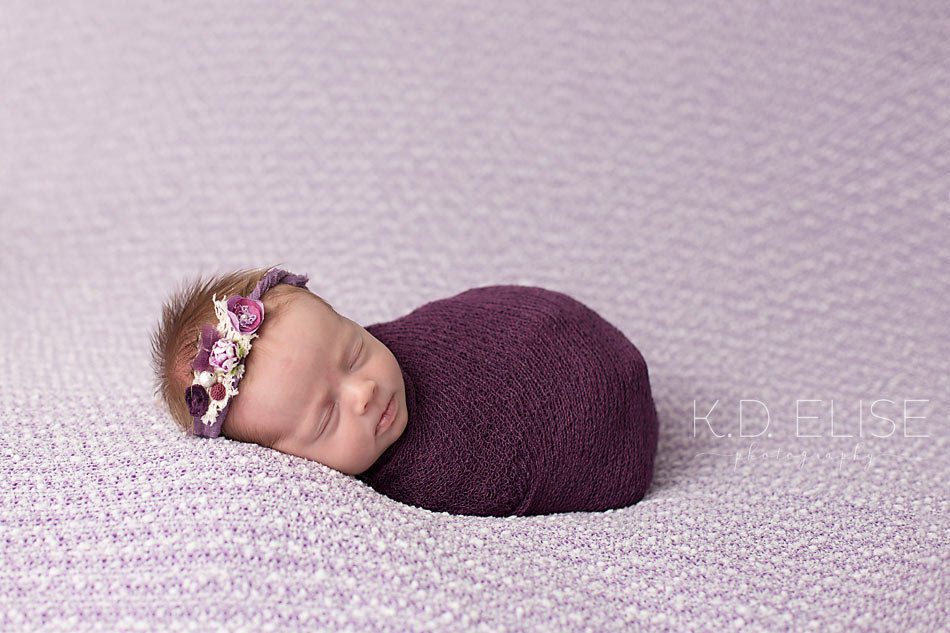 Baby girl in purple wrap and purple headband lying on a purple backdrop by Pueblo photographer K.D. Elise Photography.