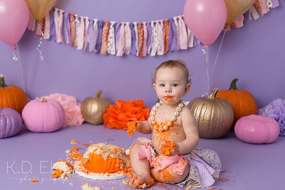 Cake smash photo shoot in a fun pink, gold, purple and orange color scheme with a pumpkin theme.