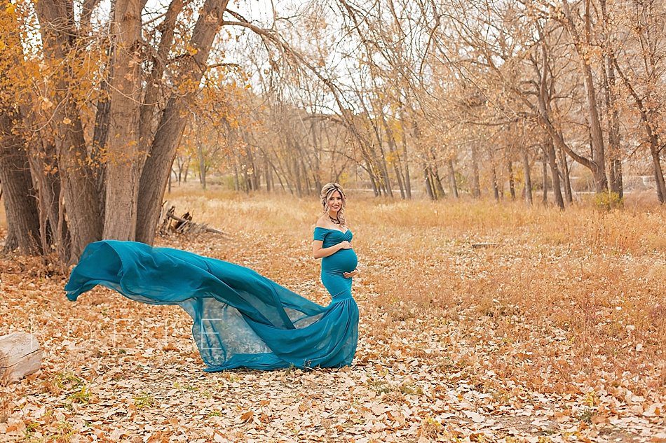 Expecting mother in a teal gown stands in a leaf covered field. Maternity portraits by Pueblo maternity photographer K.D. Elise Photography.