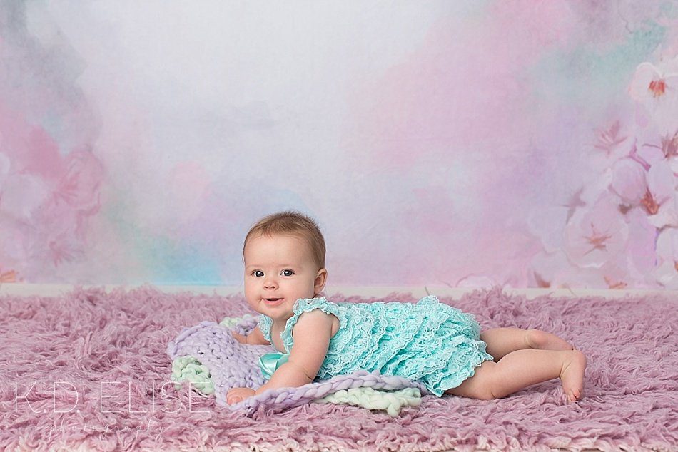 6 month baby photos of baby girl in mint romper in front of pink and purple floral backdrop. By K.D. Elise Photography, a Pueblo photographer.