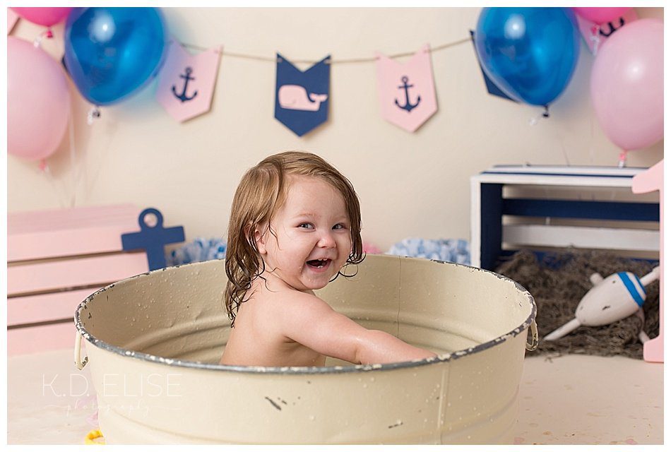 Smiling one year old baby girl in a metal wash basin after cake smash session with Colorado Springs photographer K.D. Elise Photography.