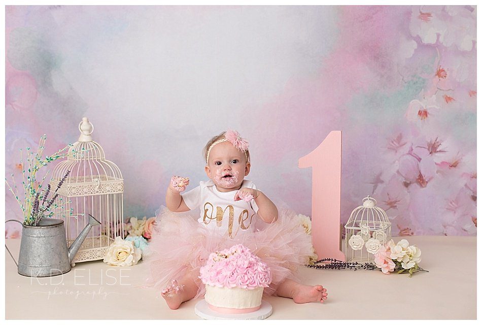 Cake smash photo of baby girl in front of floral backdrop with flowers and metal bird cages.