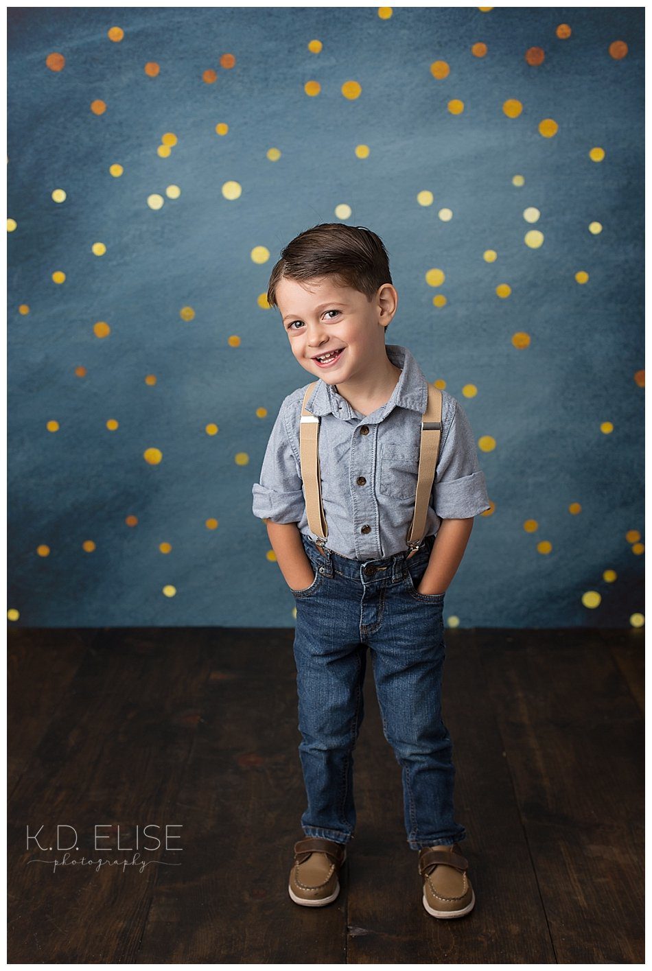 Smiling three year old boy in jeans and suspenders.
