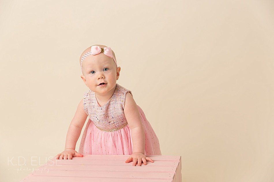 Baby girl in a pink dress standing against a pink crate before her candy themed cake smash with Colorado Springs photographer K.D. Elise Photography.