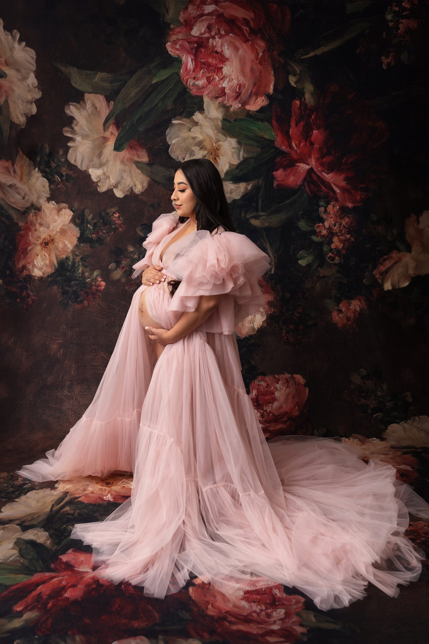 Pregnant mom in a flowing pink dress, holding her stomach in front of a floral backdrop.