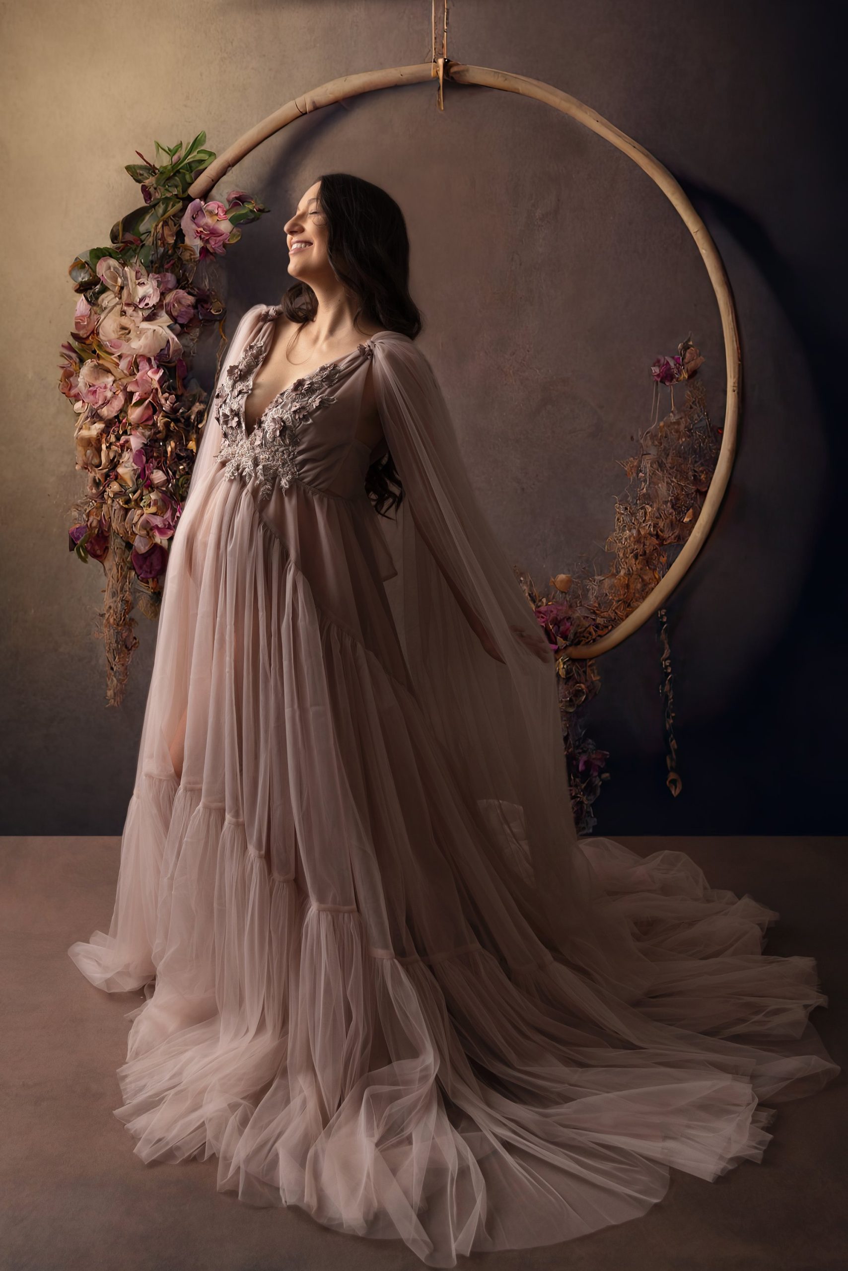 Maternity photos of a pregnant mom in a mauve gown, standing in front of a floral hoop.