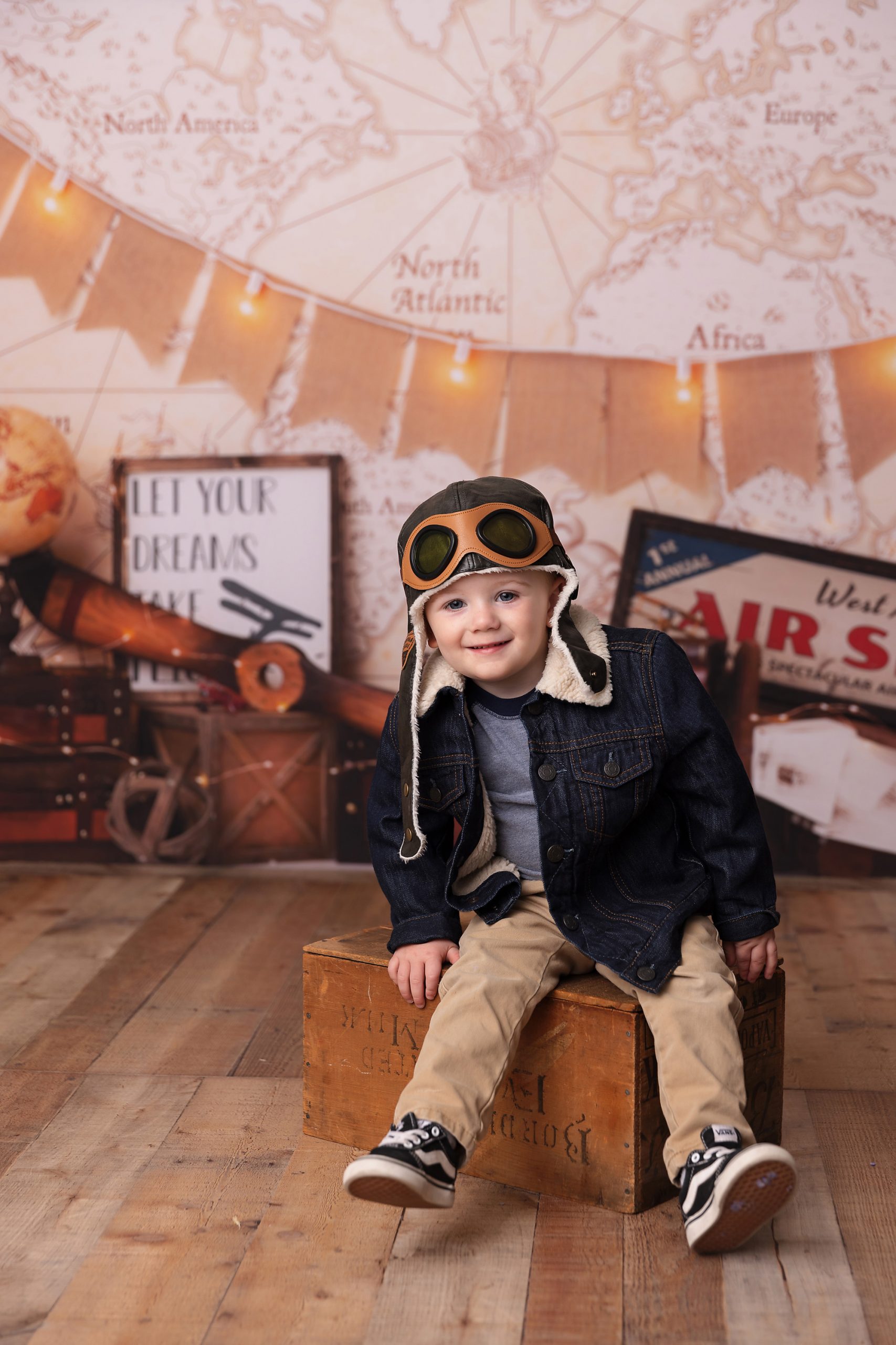 Boy in aviator jacket and hat sitting on a wooden crate in front of an airplane themed backdrop.