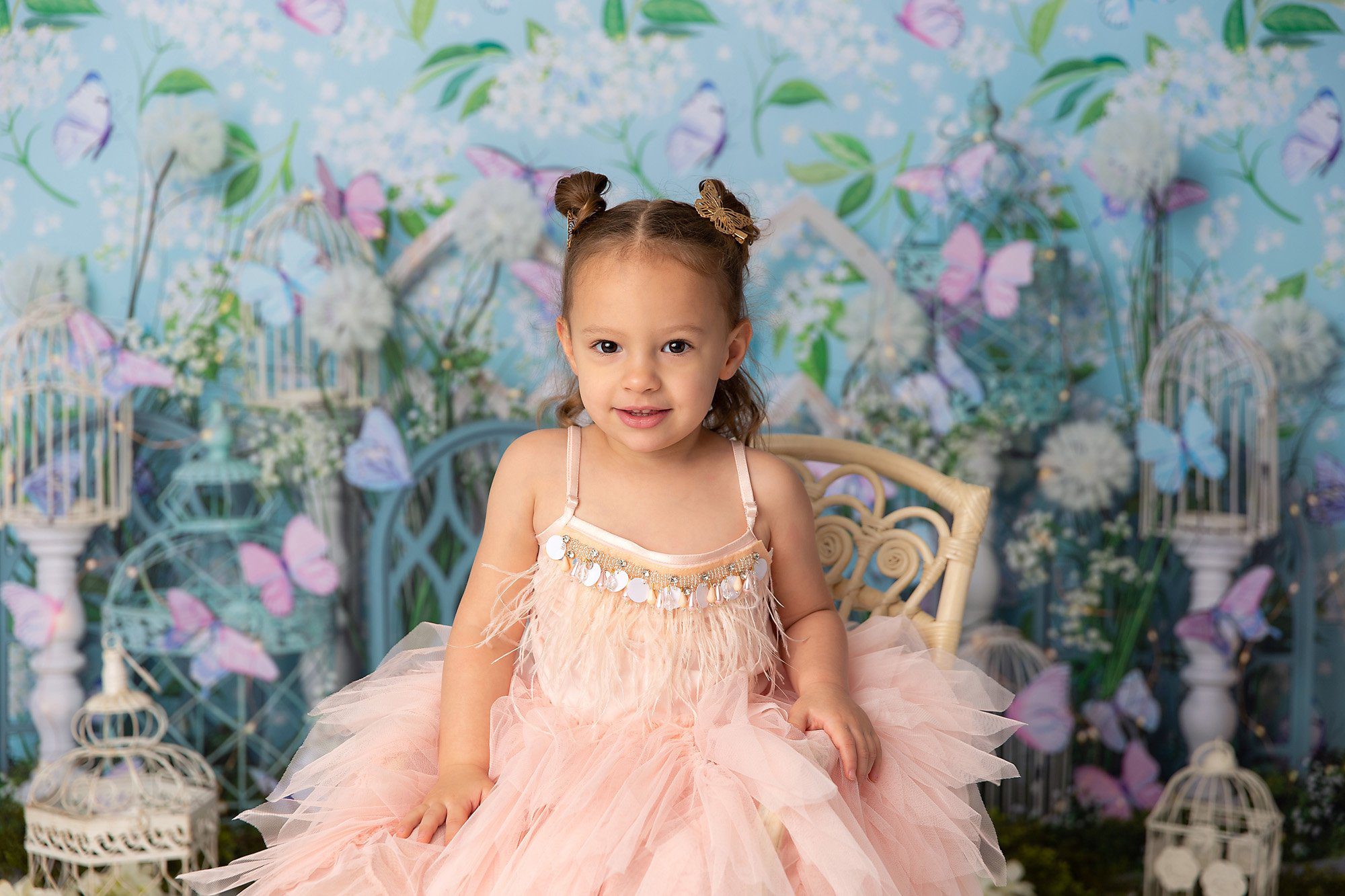 Girl in fluffy peach dress sitting in a wicker chair in front of a blue and green butterfly backdrop.