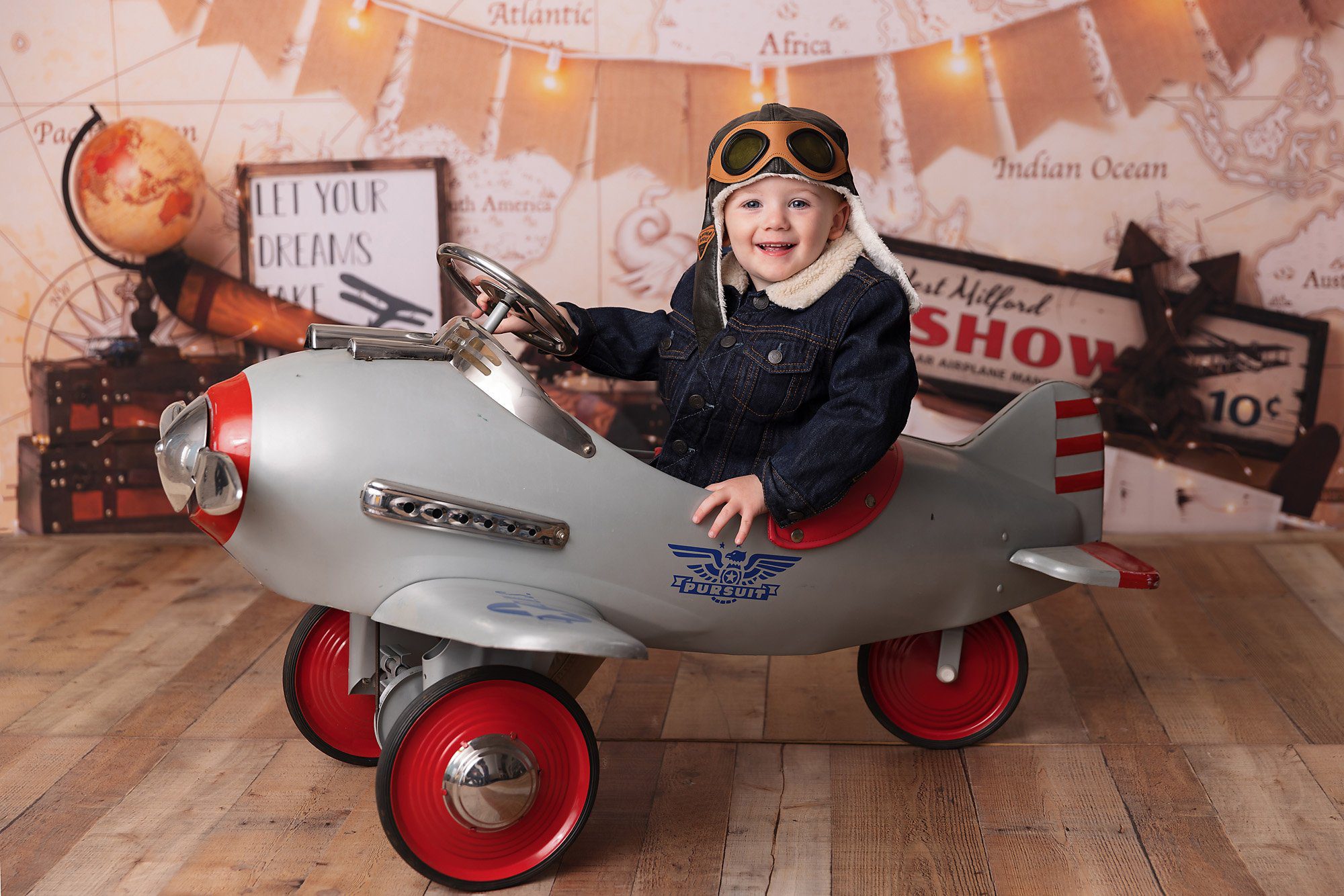 Boy wearing an aviator jacket and cap sitting in an airplane pedal car in front of a plane themed backdrop.