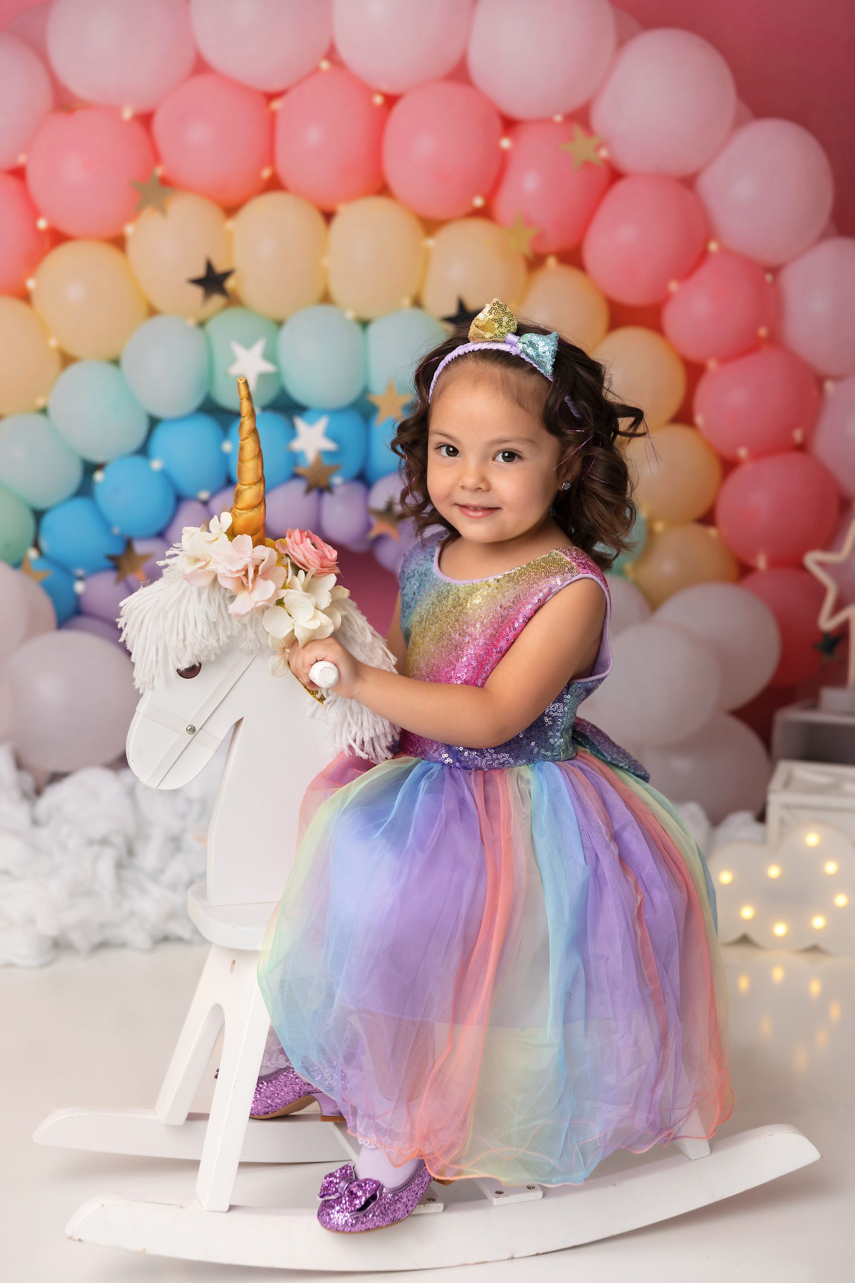Young girl in a rainbow dress on a white unicorn rocking horse. The backdrop is a pastel rainbow balloon garland.