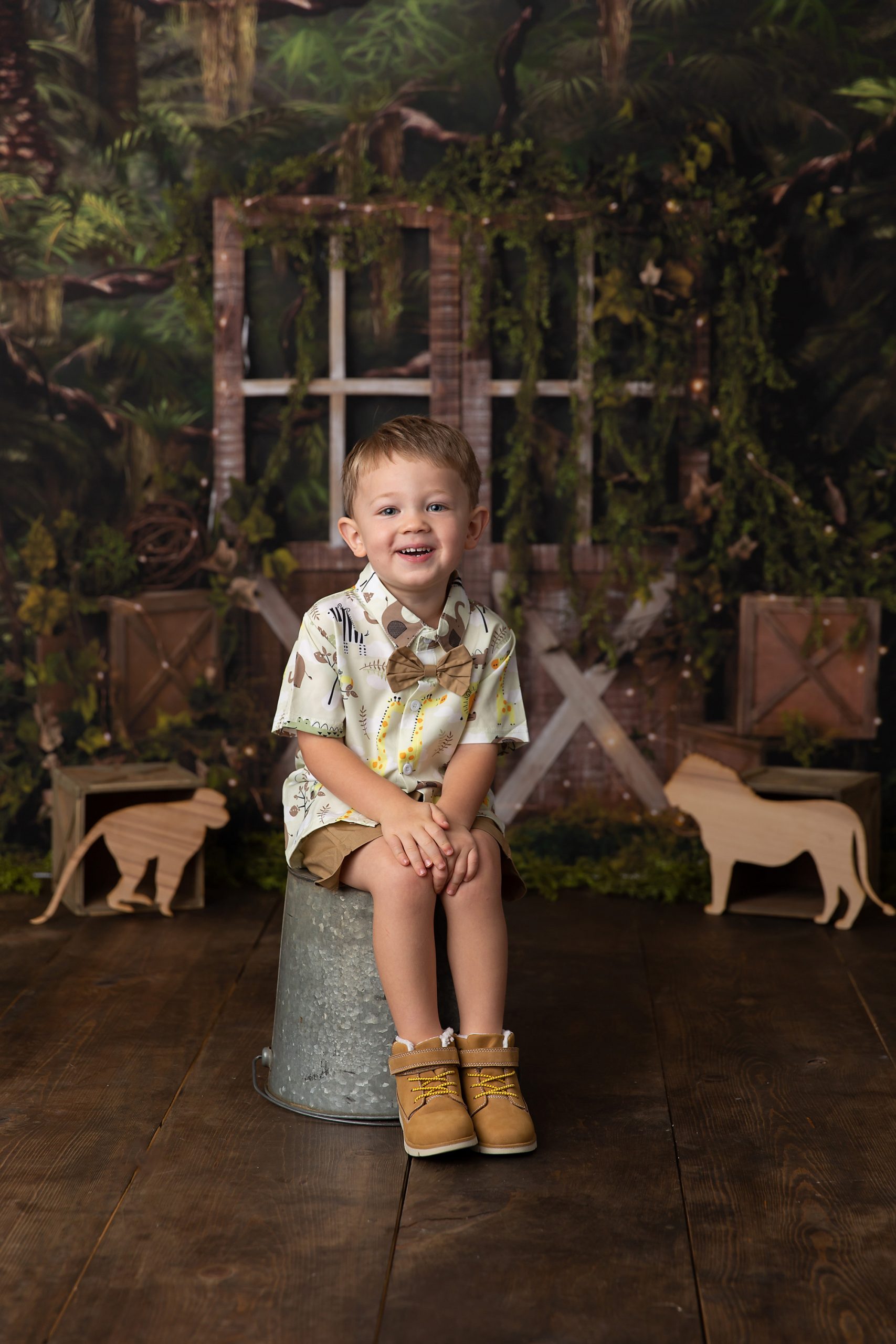 Safari themed photo of boy sitting on a metal bucket. In the background there are leaves, a wooden door, and wooden cut out safari animals.