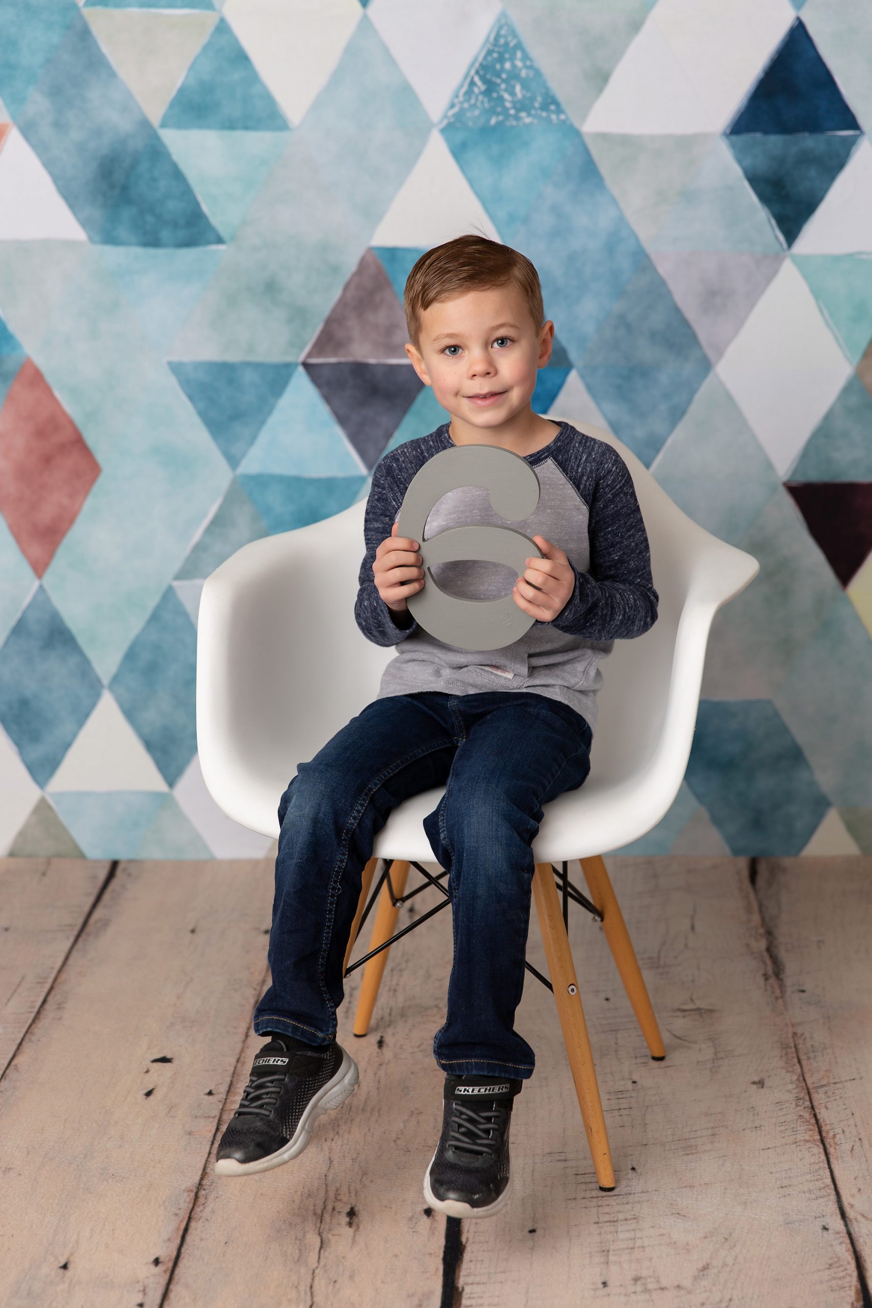 Six year old boy sitting in a white chair in front of a blue and white geometric backdrop.
