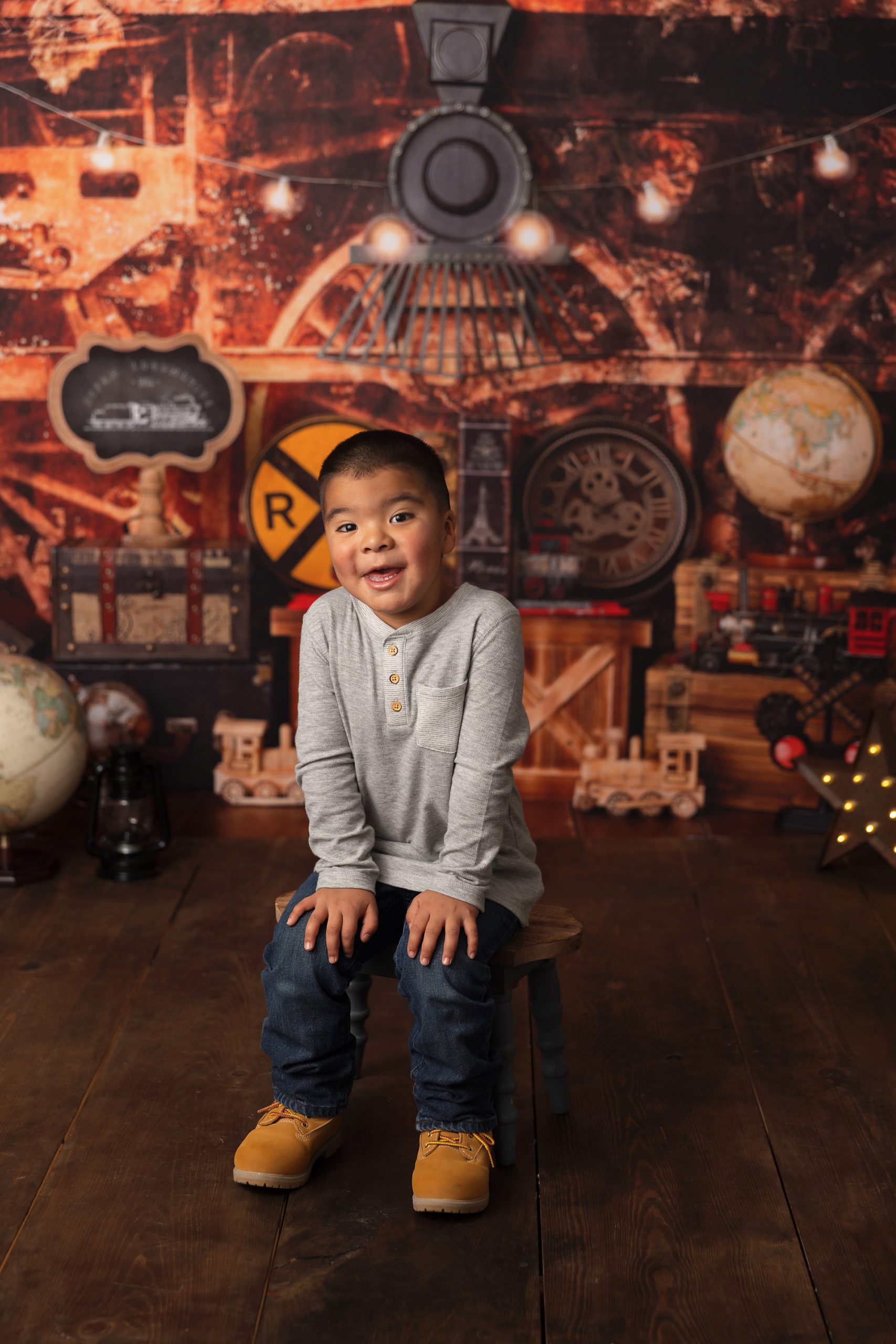 Boy in grey shirt and blue jeans sitting in front of train themed photo backdrop.