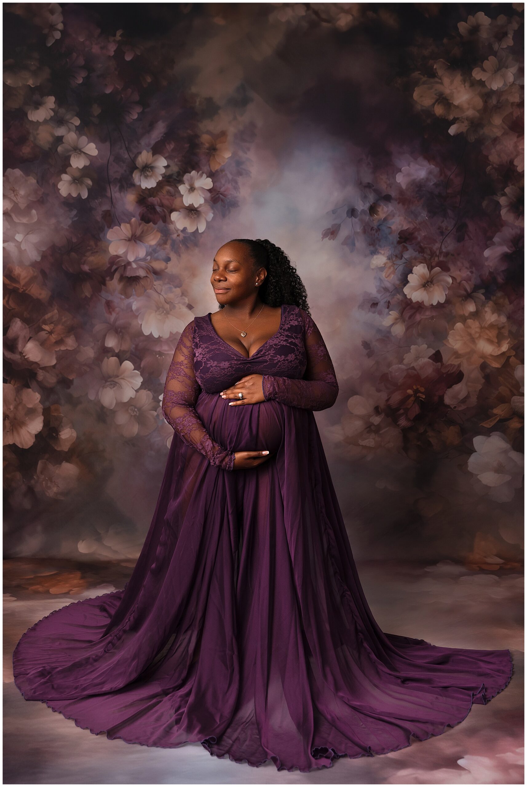 Pregnant mom in a dark purple dress. She is cradling her belly and looking off to the side. Behind her is a purple and cream floral backdrop. Photography by K.D. Elise Photography, Pueblo CO.
