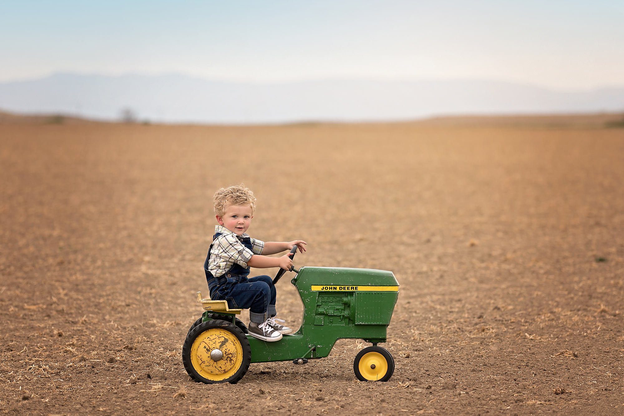 Little boy on a John Deere pedal car tractor sitting in a field in Pueblo, CO. Photo by Pueblo Colorado Springs children's photographer K.D. Elise Photography.