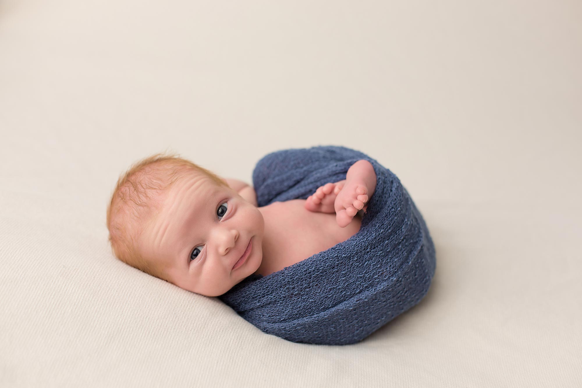 Newborn baby boy wrapped in blue, smiling at the camera. Photo by Pueblo Colorado Springs newborn photographer K.D. Elise Photography.