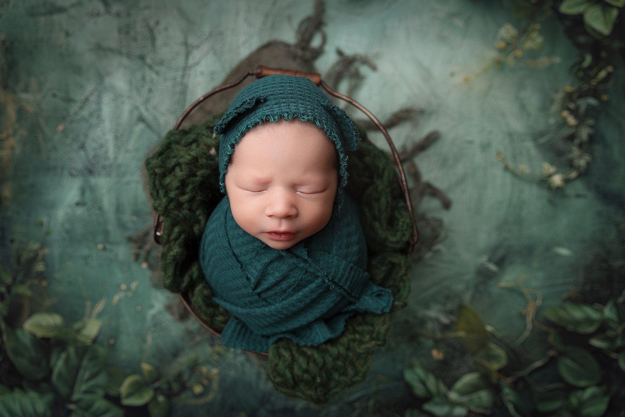 Newborn baby boy wrapped in green and wearing a green bear bonnet. He is laying in a bucket and behind him is a green backdrop.