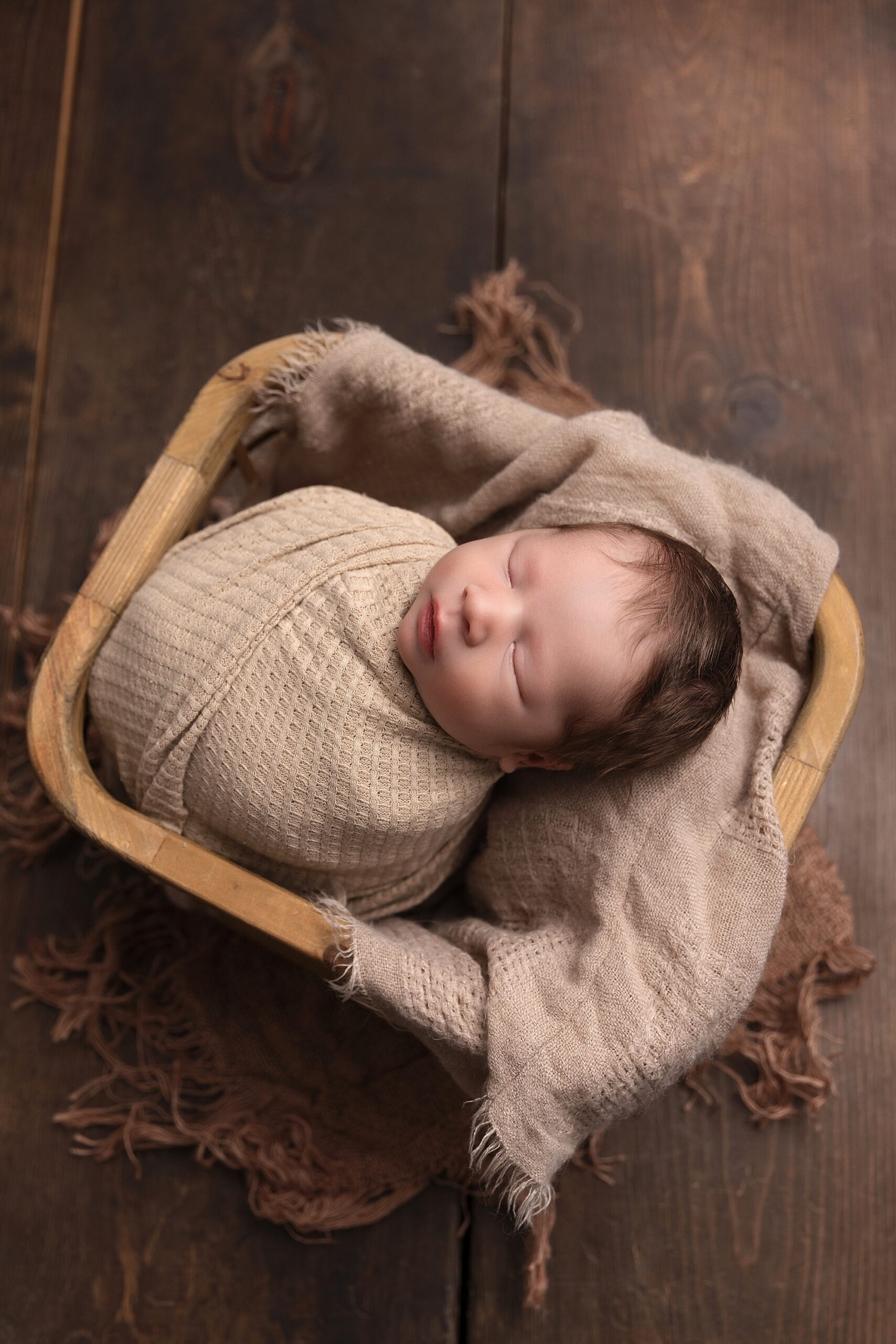 Newborn baby boy wrapped in a tan wrap laying in a wooden basket.