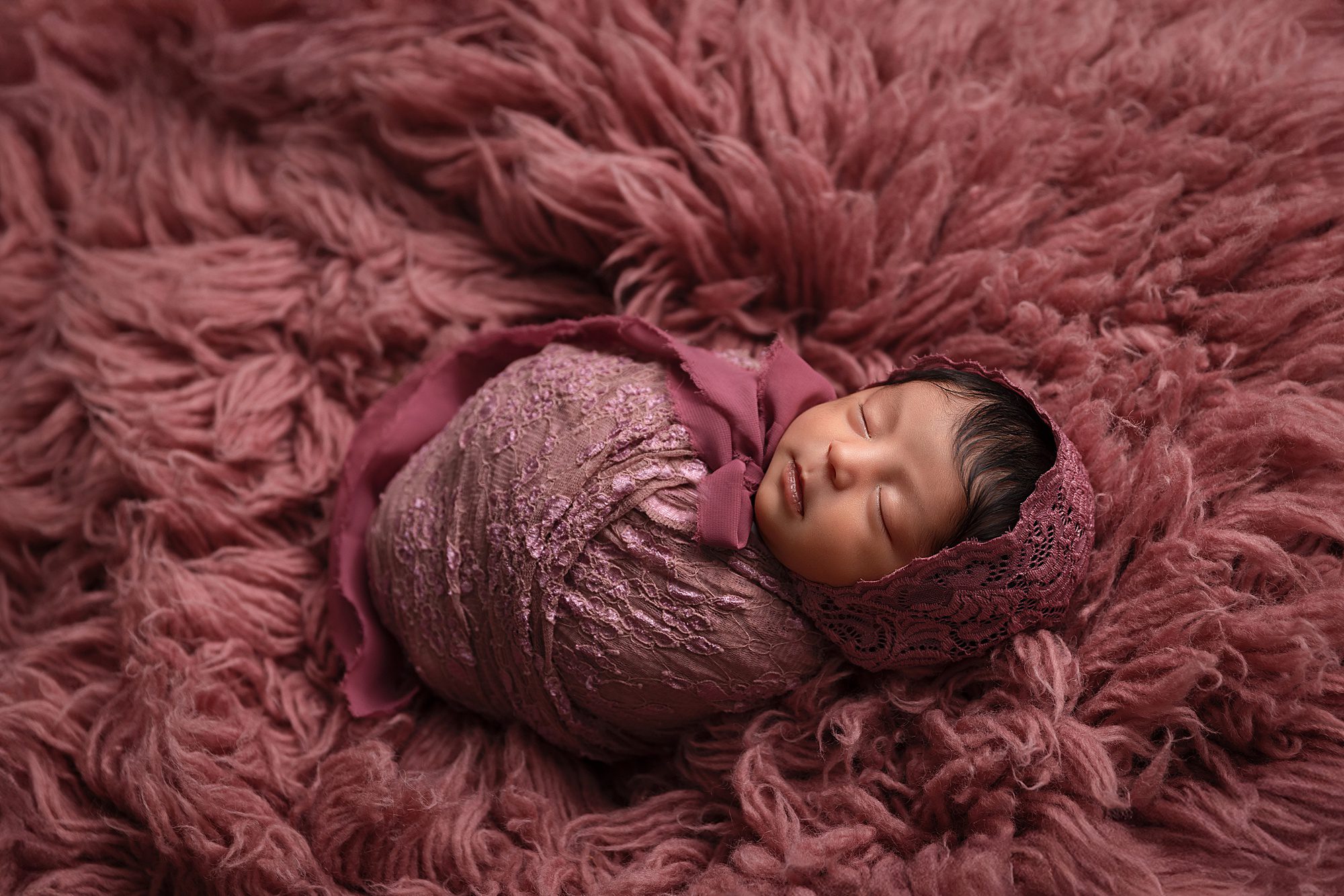 Newborn baby girl wrapped in pink, wearing a pink bonnet. She is laying on pink fur.