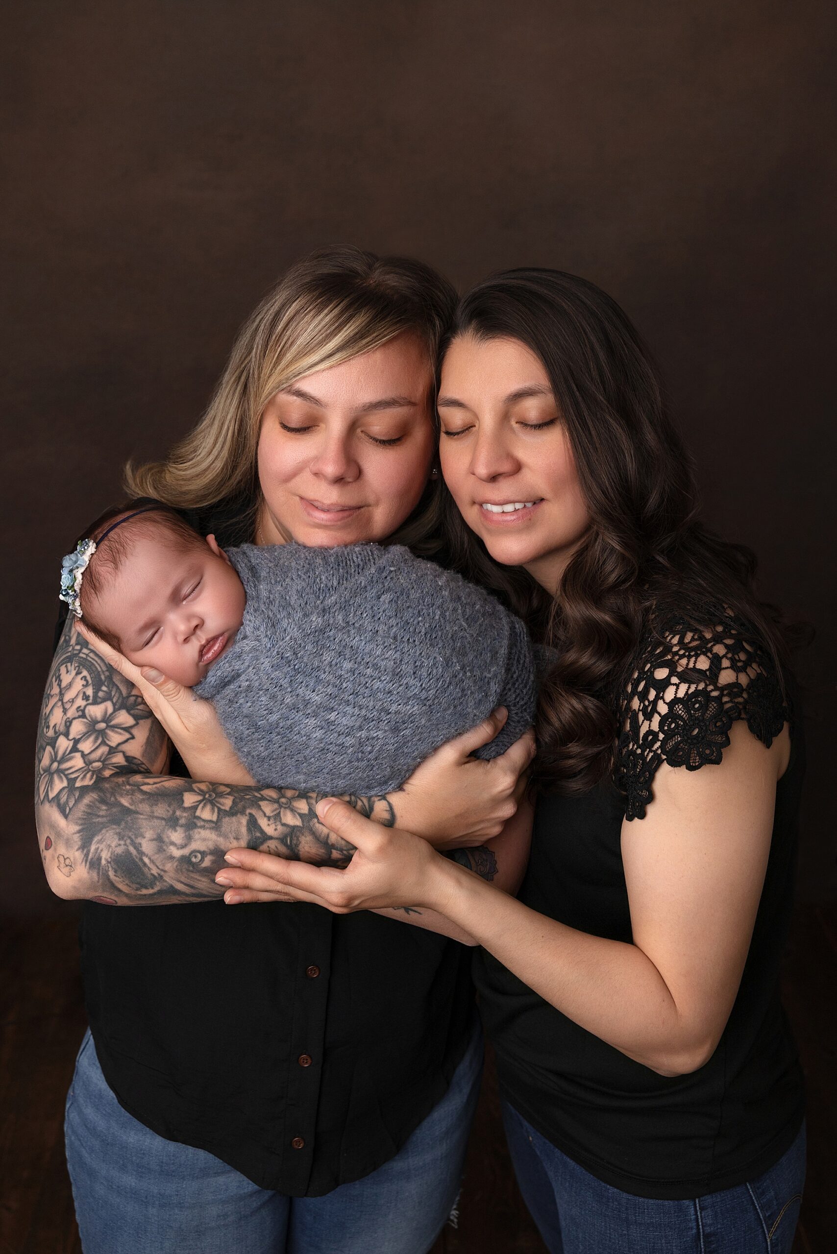 Newborn baby girl in blue wrap being held by her moms. They are wearing black shirts. Behind them is a brown backdrop.