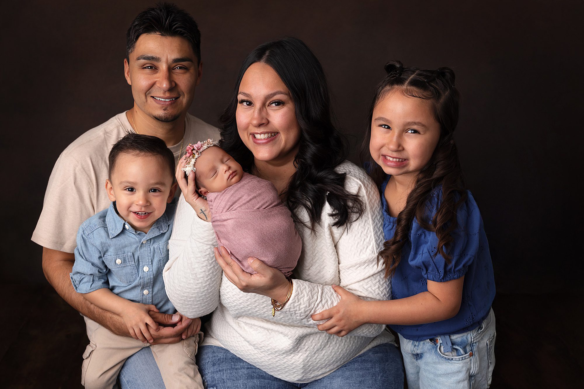 Family newborn photo. Newborn baby girl wrapped in pink being held by her mom. Surrounding mom is dad, big brother, and big sister.