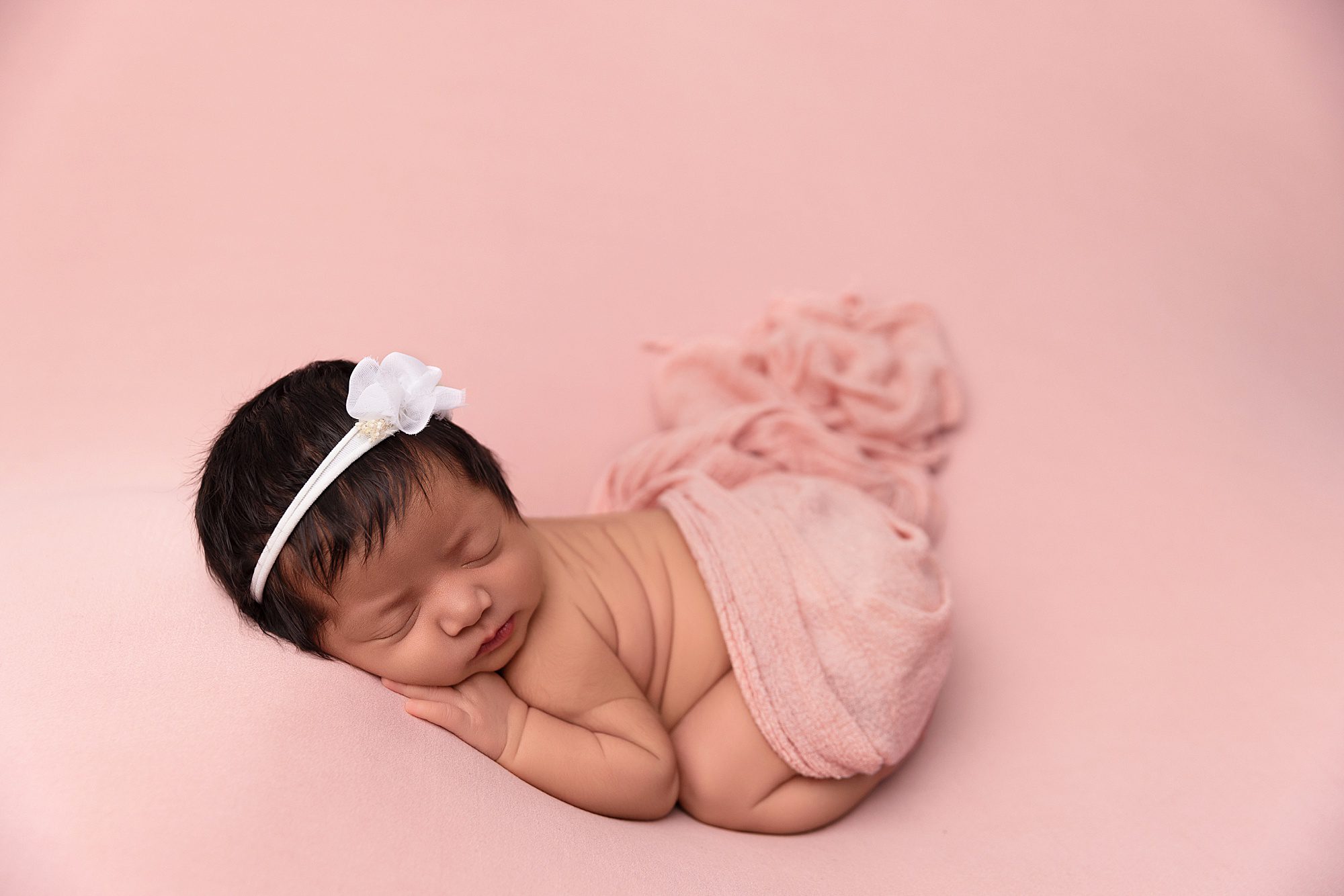 Newborn baby girl laying on a pink backdrop. She is wearing a white headband.
