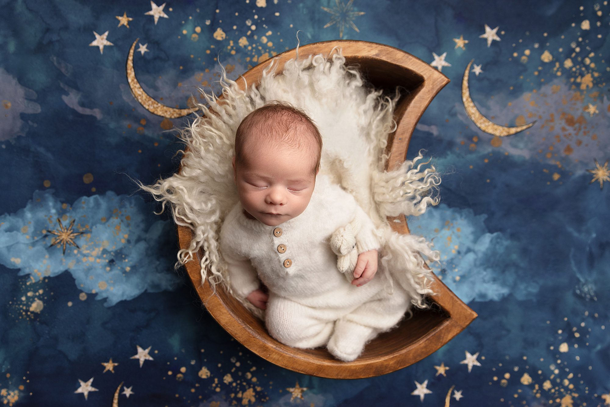 Newborn baby boy in a white sleeper. He is laying in a moon shaped bowl. In his arm is a small white teddy bear. The backdrop behind is has moons and stars.