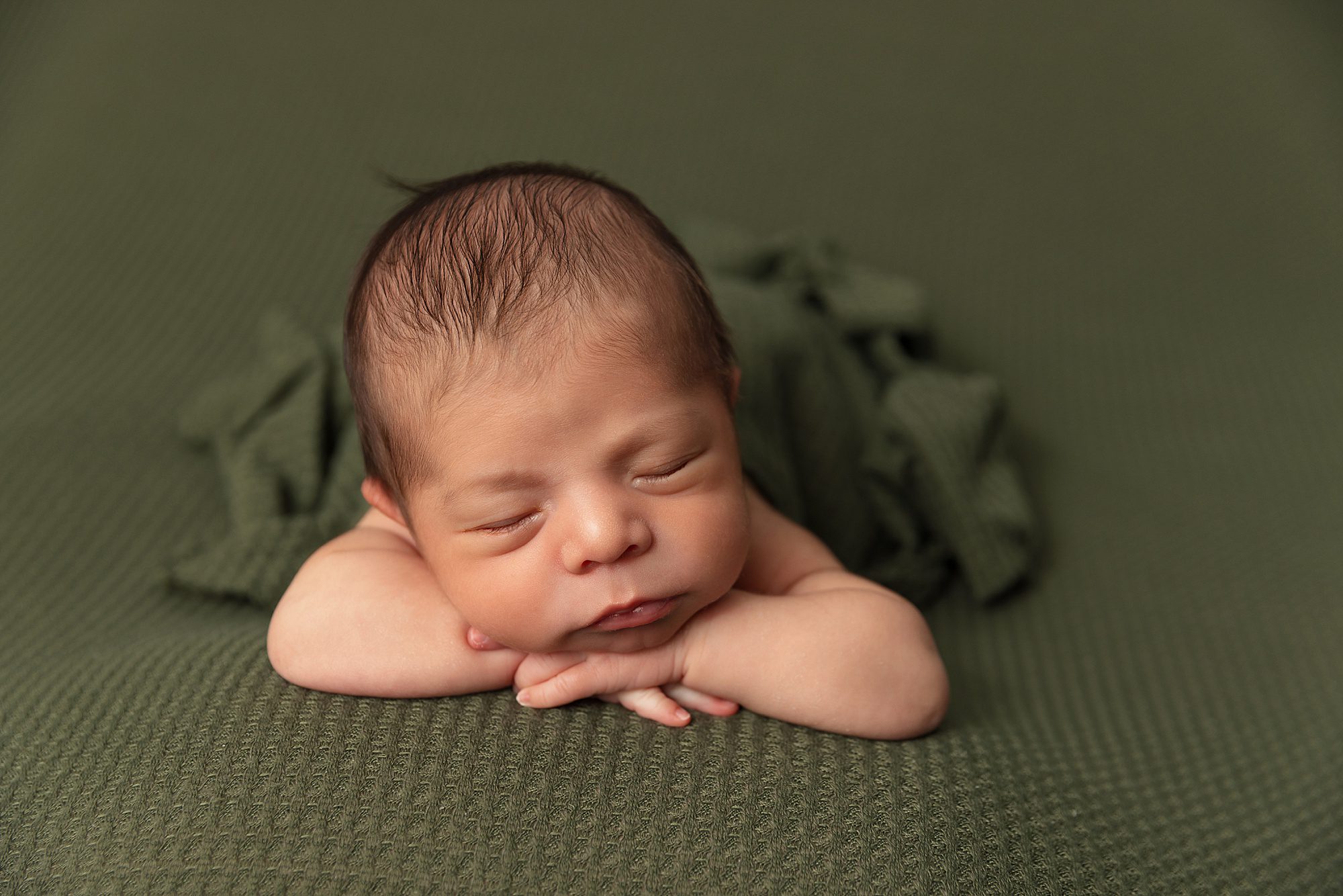 Newborn baby boy laying on a green blanket. A green wrapped is tucked around his body.