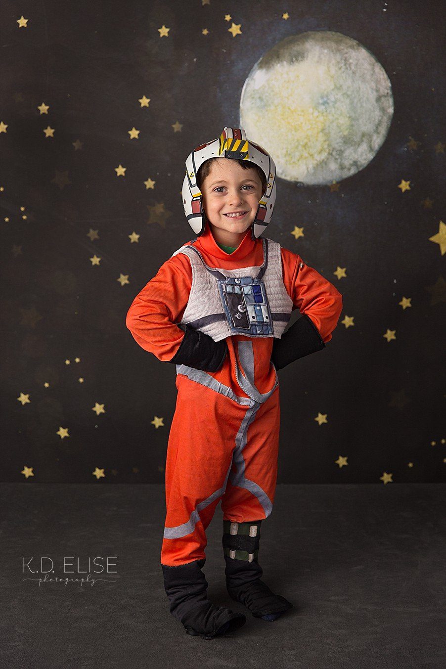 Little boy in a flight suit costume in front of a space themed backdrop. 