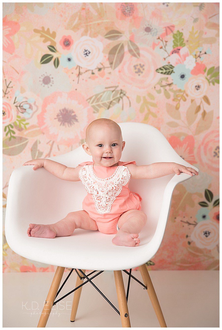 Baby girl in a white chair sitting in front of a floral backdrop.