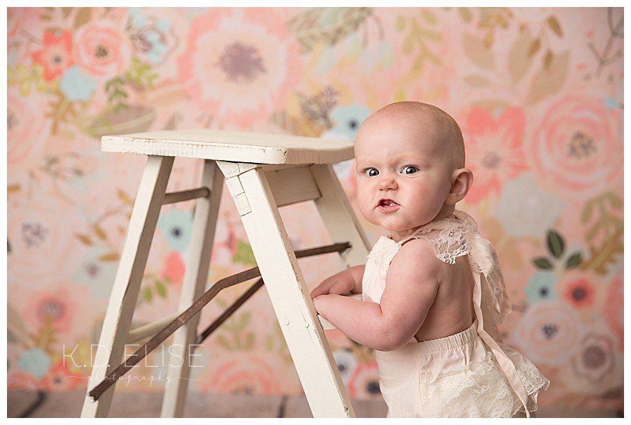 9 month milestone session of baby girl standing against a ladder in front of a floral backdrop.