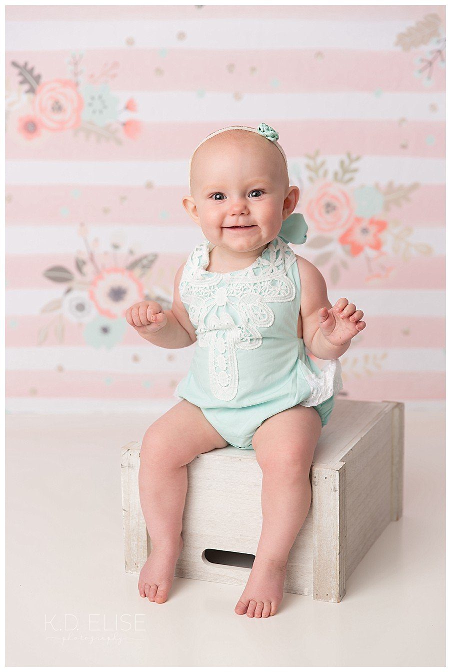 Baby girl sitting on a crate in front of a pink and white stripped backdrop. 