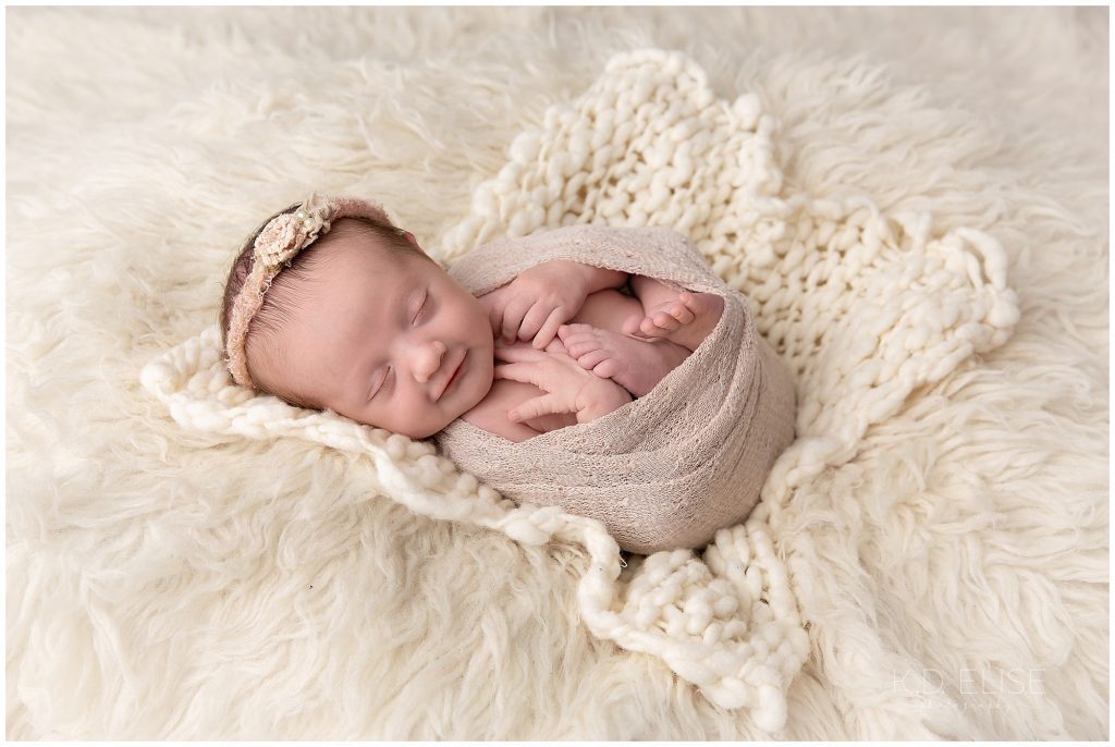 Newborn baby girl wrapped in pink, laying on a cream fur.